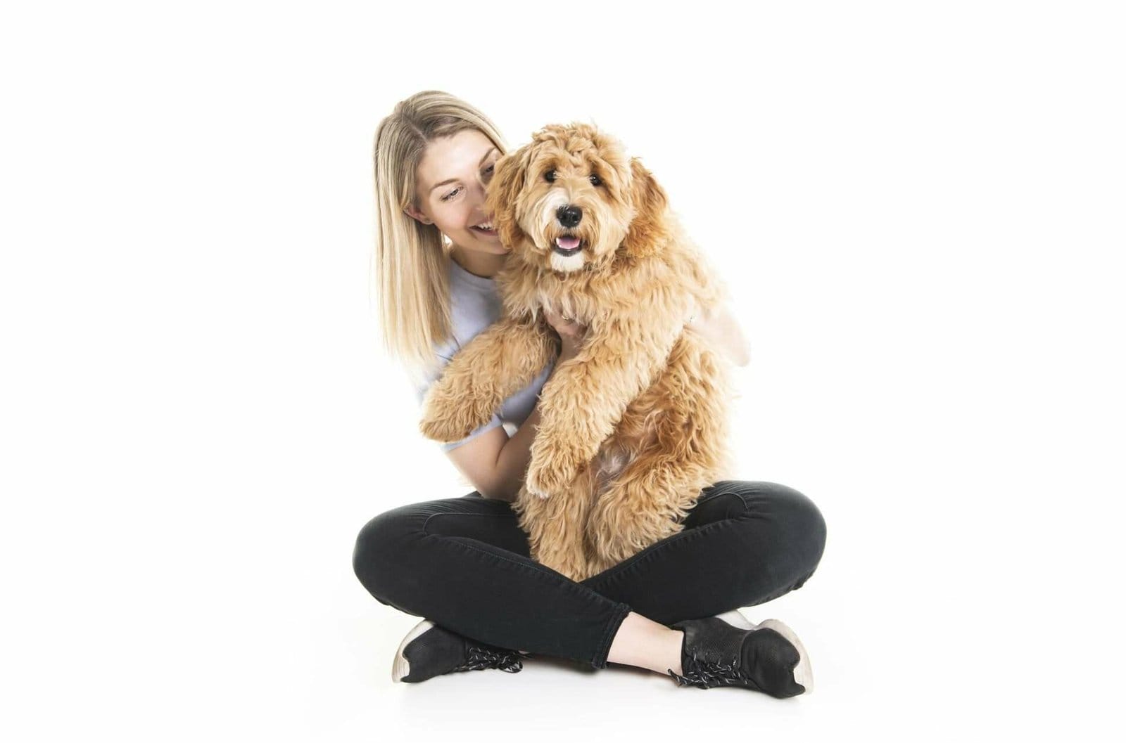 a woman sitting on the floor smiling, and holding a mini Goldendoodle dog on her lap and hands